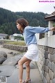 A woman in a blue sweater and white skirt leaning against a stone wall.